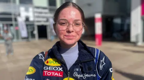Simon Thake A young woman with hair tied back and glasses stands in the sunshine wearing a navy jacket with colourful patches