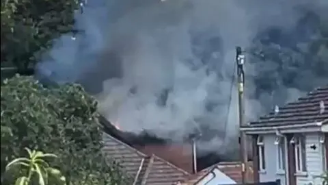 Jamie Somerville A slightly blurry picture mostly showing thick smoke coming from the top of a house. Other homes around and some tree branches to the left