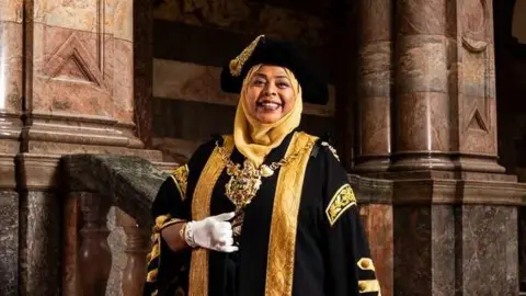 A woman is wearing a gold hijab and over the top a black tricorne hat with gold braid. She is wearing black and gold ceremonial Lord Mayor robes and white gloves and smiling. She is stood on a marble staircase in Sheffield town hall.