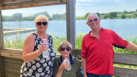 Shariqua Ahmed/ BBC Sharon, a woman wearing a balck and white floral top standing next toMrs Allen, who is a woman in a green top and Bill Allen standing next to her, a man in a red top.