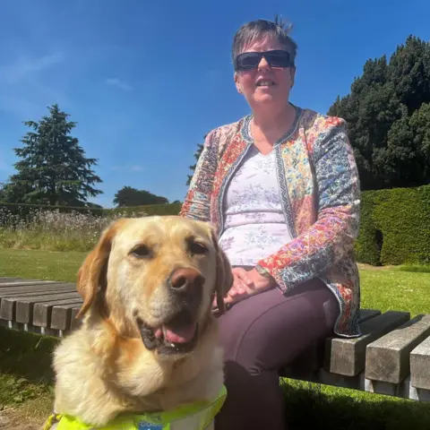 Sylvia Paton Sylvia Paton senta-se num dia de verão em um banco de madeira ao sol com céu azul. Ela está usando óculos escuros, pois a luz forte afeta sua condição ocular. Ela tem sua cadela labradora loira, Kate, em primeiro plano.