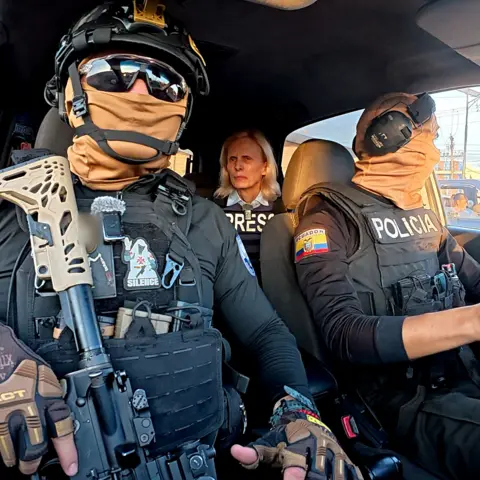 Lee Durant/BBC Two Ecuadoran police officers in dark sunglasses, helmets and balaclavas - which entirely obscure their faces - sit in a car in front of the BBC's Orla Guerin, wearing a flak jacket with the word "PRESS" on it. 