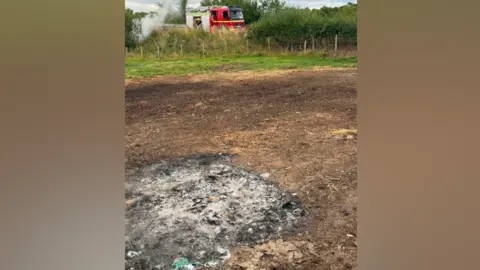 A patch of land which has been scorched and there is a black patch of ashes. It is on countryside land and in the distance there is a Cumbria Fire and Rescue appliance which a firefighter is exiting from. 