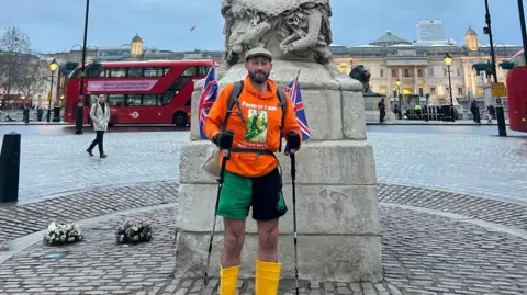 Sam Stables A man in an orange jumper, green and black shorts and yellow boots standing in front of a statue. Behind him is a red double decker bus and a person walking. 