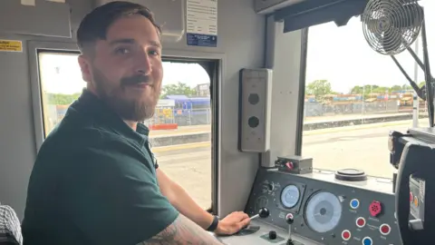 Ben Warner smiles at the camera while sitting in a drivers cab for a train. Lots of dials and leavers can be seen. He wears a green polo shirt.