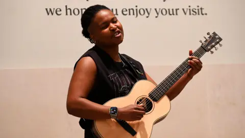 Time Out / Jake Darling Sherika Sherard, a woman with short dark hair and wearing a black vest-type top, is singing and playing a guitar.