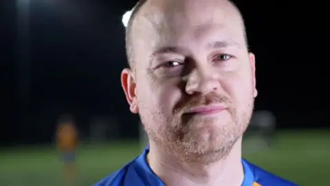 Freddie Kizis-Stevens is wearing a yellow and blue football shirt, with a Sands United crest. He is standing on a football pitch.