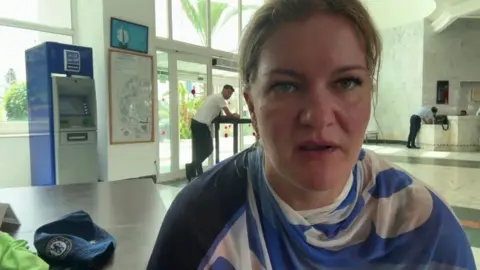 Natasha Dovbijenko A woman has taken a selfie in an airport. Behind her are a few security staff and safety barriers. There is also an ATM in the background. 