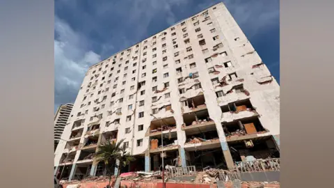 Mark Leeson A large block of flats that has been hit my an earthquake. Is has pieces of the building missing, with some parts having the flats and rooms completely exposed. There are piles of rubble underneath it.