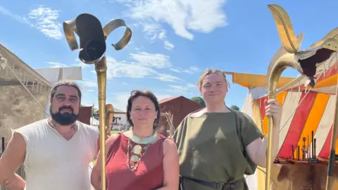 A man and two women pose for a photo with two ancient Celtic war horns. The horns are cold and have a big opening in the shape of an animal's mouth. They are tall, ending at least a head height above the people. A few tents and marquees can be seen in the background.