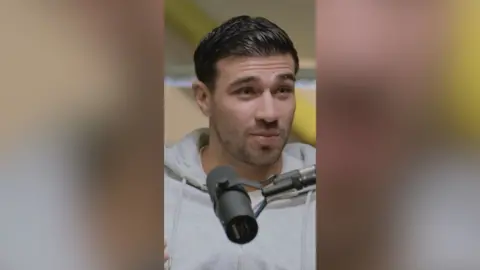 A man with slicked back dark hair and wearing a light grey hoodie speaks into a microphone.