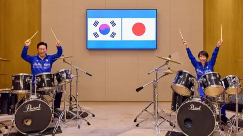 Lee Jae Myung and Sanae Takaichi sitting side by side with separate drum sets. They are raising their drumsticks in the air and smiling.