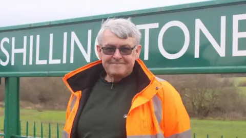 A smiling Gavin Collins standing in front of one of Shillingstone's green station signs. Behind him is countryside. He is wearing glasses and an orange hi-viz coat.
