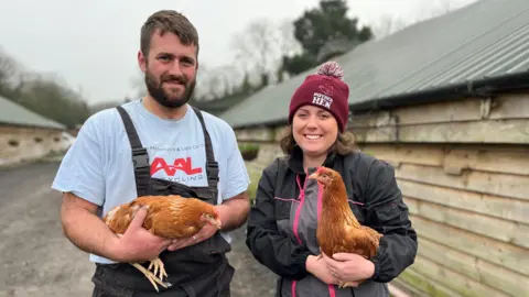 BBC A man with a beard in a grey T-shirt and black overalls holds a chicken next to a woman with a maroon hat which says "mother hen" on it. She is also holding a chicken. 
