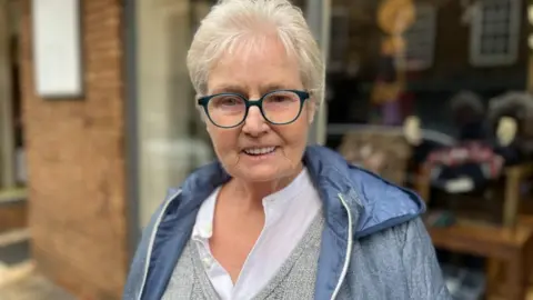 BBC A picture of Maggie Clark, who is wearing glasses, a shirt, vest and jacket.