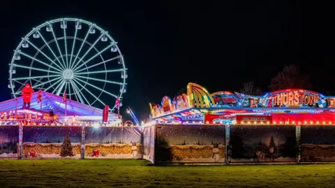 Bronson Thurston A view of a fairground from a distance at night. There are stalls in front with a ferris wheel lit up in the background.