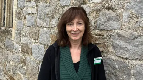 Dr Ros Cleal is wearing a dark green jumper and a black blazer with a name tag. She has brown hair and is smiling at the camera.