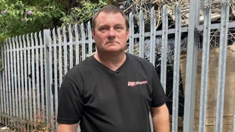 BBC/PHIL BODMER Man in a black tee shirt stands next to a metal fence behind which is an illegal waste dump