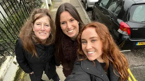 Lucy and daughters Izobel and Eliza standing on the pavement, in a selfie taken by Eliza. Lucy has long light brown hair and wears a black winter coat. Izobel has long dark brown hair and wears a dark red fluffy coat. Eliza has long red hair and wears a black jacket.