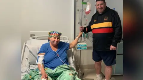 Jenny in hospital bed during stem cell transplant with Colin standing next to her bed holding her hand. There is a drip stand between them.