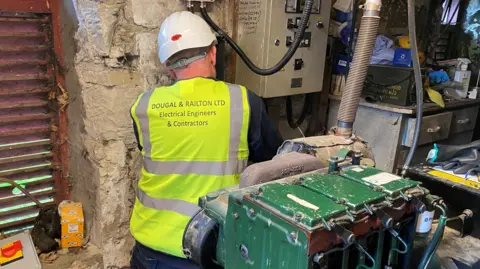 An engineer wearing a hi-vis jacket is working next to an electricity meter with a green generator behind him 