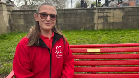 BBC/Chloe Hughes Zoe has brown hair and is wearing tinted glasses. She is wearing a red T-shirt and a red jacket. The jacket has a white British Heart Foundation logo on its breast. She is sitting on a red bench with a gold plaque and there is green grass and a stone wall behind it