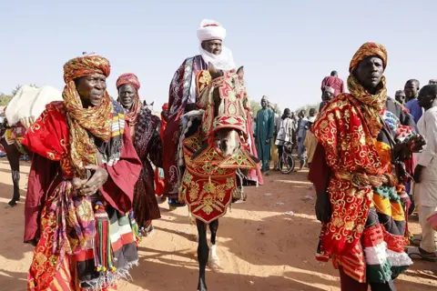 Anadolu via Getty Images A man in traditional attire and turban is on a horse which is dressed in embroidered red cloth. Two men, in matching clothes are in front leading the procession.