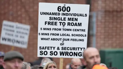 Eddie Mitchell A sign reading "600 unamed individuals, single men free to roam, 5 mins from town centre, 5 mins from local schools, what about our feeling of safety & security? So so wrong!"