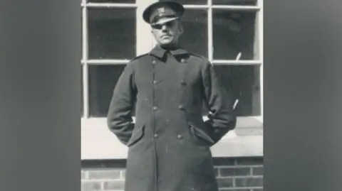 Essex Police A man standing with his hands behind his back. He is wearing a long black coat and a police hat. He is standing in front of a building.