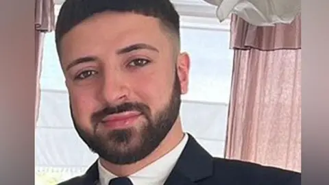 Head and shoulders photo of Kyle Clifford smiling at the camera wearing a black jacket and tie and white collared shirt.
