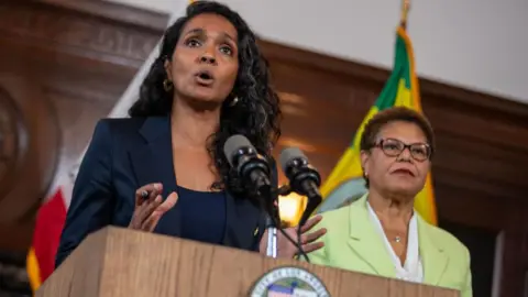 Los Angeles Times via Getty Images Nithya Raman stands at a podium and microphone with Karen Bass standing alongside her 