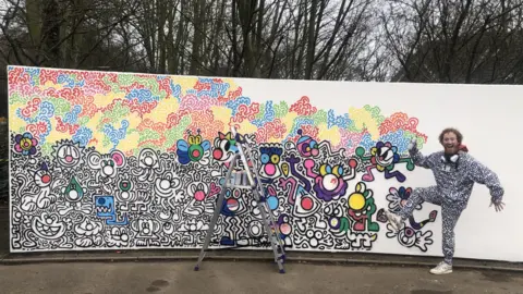 ALEXANDER STEVANOVIC/BBC Mr Doodle is standing on one leg by a mural which is mostly covered by all sorts of colours and images of smiley cartoonish looking characters. There is a ladder with paint cans on it, in front of the mural.