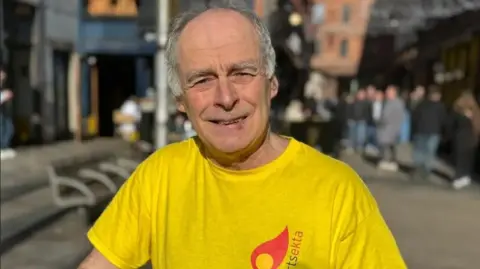 A man, Séamas Ó Labhradha pictured outside in Belfast city centre. He has grey hair and is wearing a yellow top with the arts organisation's logo Arstekta on it in red and black.
