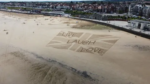 Fred Brown A large sand artwork on Bridlington beach, with a Union Jack flag and the slogan live, laugh, love, in a comic sans font. In the background are houses and hotels on Bridlington sea front.