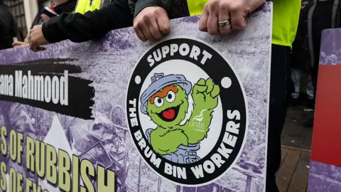 Two people in high-visibility vests grip a large purple and black banner reading 'Support the Brum Bin Workers' alongside a cartoon character in a dustbin with the Unite the Union logo. Other protesters can be seen standing behind the banner in the background.