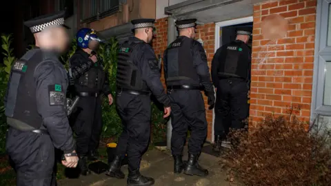 Five uniformed police officers burst through a front door in a raid