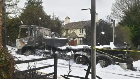 Shropshire Fire and Rescue Service The blackened shell of a lorry in a street covered in white foam, with a house in the background and a lamp post and fence in the foreground.
