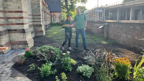 Woman wearing green T-shirt, cap and dark jeans with man wearing green T-shirt with blue jeans stand on a patch of soil behind plants, next to a paved area. There is a brick and stone church building to one side of them and a brick wall and another building to the other.