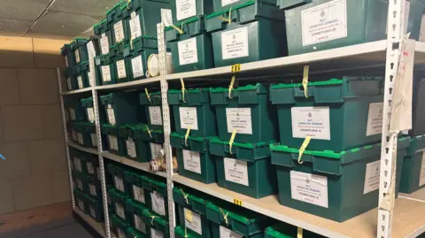 Avon and Somerset Police The inside of a storage locker at Avon and Somerset Police. It shows multiple shelving units, stacked high with approximately 50 green plastic boxes containing evidence and files related to the investigation. 