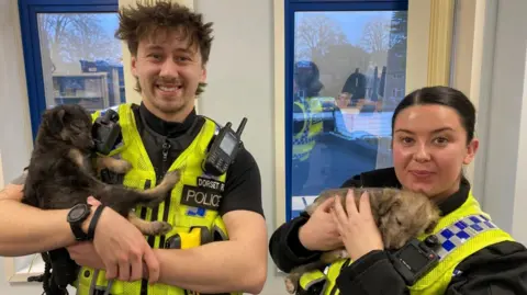 Two unnamed police officers in police jackets standing while cuddling two puppies, which are both sleeping.