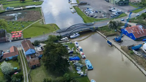 Oliv3r Drone Photography A drone image of a white cruiser wedged under a metal bridge over a river, with a marina in the distance.
