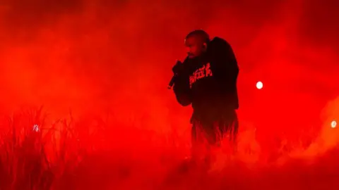 Getty Images Kanye West is performing on stage. He is hunched over with a mic in one hand, which he is holding up to his face. He is wearing a black hoodie with writing in a different language on it and black trousers. He is surrounded by straw sprouting up from the ground and clouds of smoke. There are bright red lights in the background.