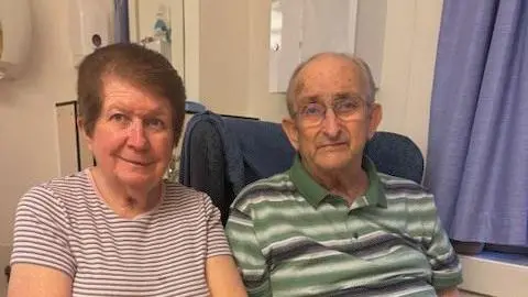 Linda Allen, sat in a stripey T-shirt and with short brown hair, and Terry, in a striped green shirt, are sat in a hospital room, with a sink and curtains behind them. 