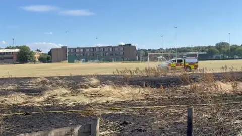 A large area of burnt grassland near a school playing field, with a fire vehicle near it.