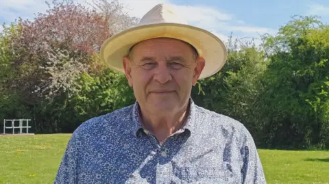 Rick Hill A man with a blue and white shirt and a hat. Grass is behind him, along with trees going from left to right. 