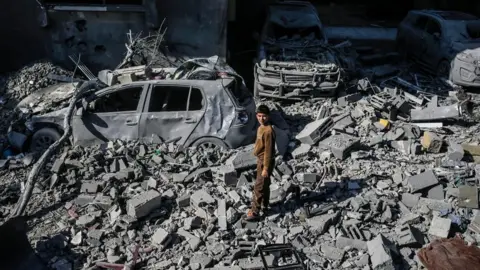 EPA A pre-teen boy stands amongst grey rubble and burned out cars, squinting up towards the camera