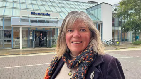 Dianna Moore pictured in front of the Riverside leisure centre in Exeter. The building is mainly built with glass panels. A sign is displayed on the glass roof with blue coloured font reading 'Riverside'. Diana is in front of the building. She is smiling ,wearing a cream roll neck jumper, a navy blue scarf with an orange and yellow thread pattern running through it. She is wearing a purple blazer on top of the jumper.