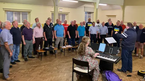 Male voice choir standing in a semi circle singing in a room which has cream walls and parquet flooring. In front of them with his back to the camera is the conductor and to his left is a female pianist who is wearing a floral dress.