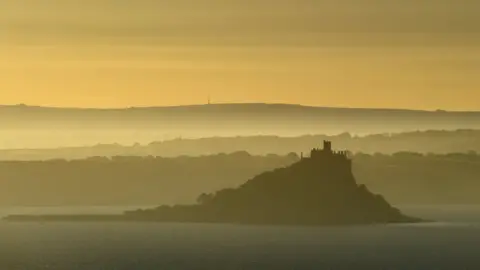 Reuters St Michael's Mount is seen shortly after sunrise. The sky is yellow/orange and the mount is silhouetted against the backdrop.