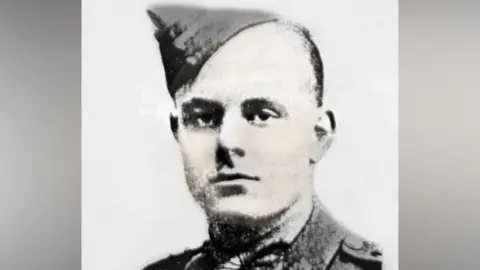 A black and white image of a man with short dark hair, wearing a naval uniform hat. He appears against a whitebackground.